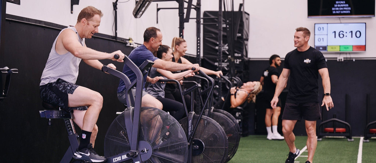 Group of people using stationary bikes in a gym with a coach observing.